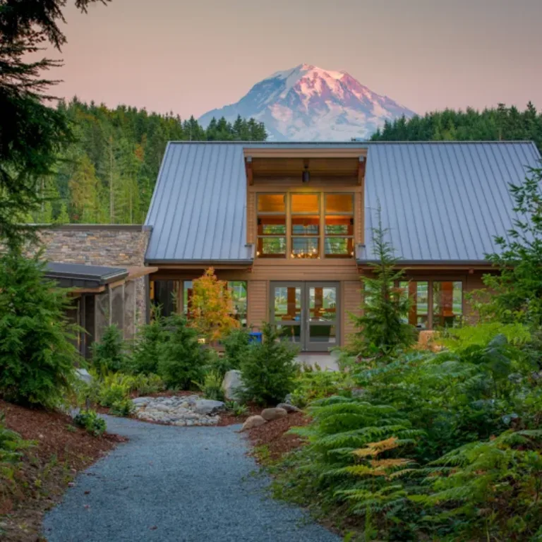 A modern timber house with a steep metal roof and large windows sits amid lush evergreens, a gravel path leading to the entrance, with a snow-capped mountain glowing at sunset in the background.