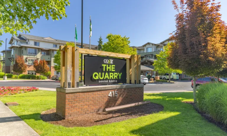 Entrance sign for Cogir The Quarry Senior Living at 415, with landscaped lawns, flowers, and the multi‑story community buildings in the background on a sunny day.