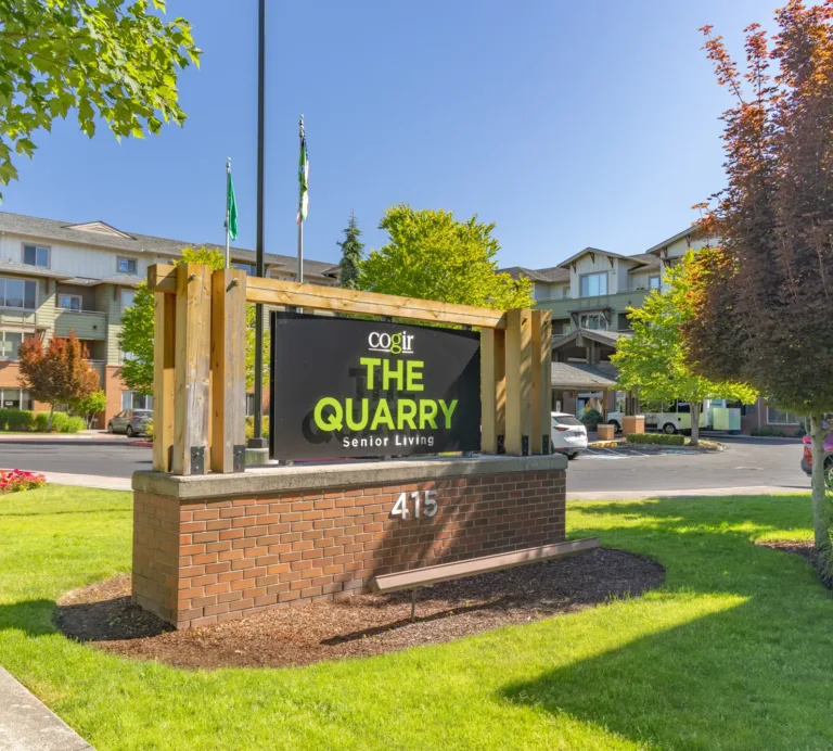 Entrance sign for Cogir The Quarry Senior Living at 415, with landscaped lawns, flowers, and the multi‑story community buildings in the background on a sunny day.