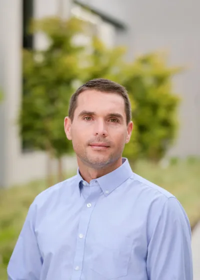 Headshot of Tony Chenier, a person with short brown hair wearing a light blue button-down shirt, posed outdoors with greenery and a blurred modern building in the background.