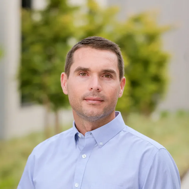 Headshot of Tony Chenier, a person with short brown hair wearing a light blue button-down shirt, posed outdoors with greenery and a blurred modern building in the background.