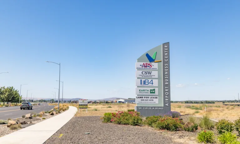 Roadside monument sign for the Airport Business Center near Tri-Cities Airport listing APS Advanced Protection Services, CSW Construction Services of Washington, IB4, Earth Solutions, and Land for Lease, set against an open landscape and clear blue sky.