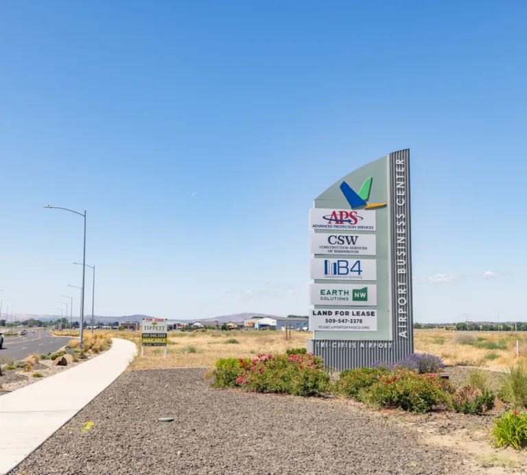 Roadside monument sign for the Airport Business Center near Tri-Cities Airport listing APS Advanced Protection Services, CSW Construction Services of Washington, IB4, Earth Solutions, and Land for Lease, set against an open landscape and clear blue sky.