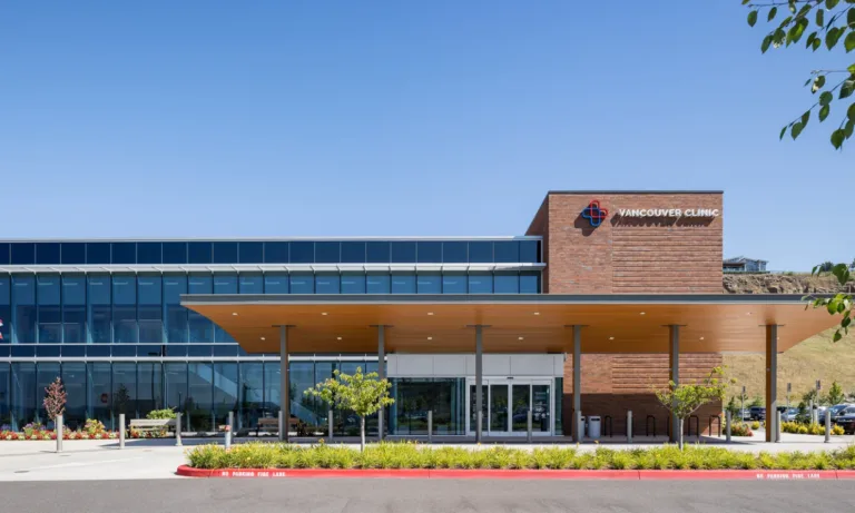 Front view of Vancouver Clinic’s modern building with glass facade and brick sign, a large covered entrance, landscaped grounds, and flags under a clear blue sky.