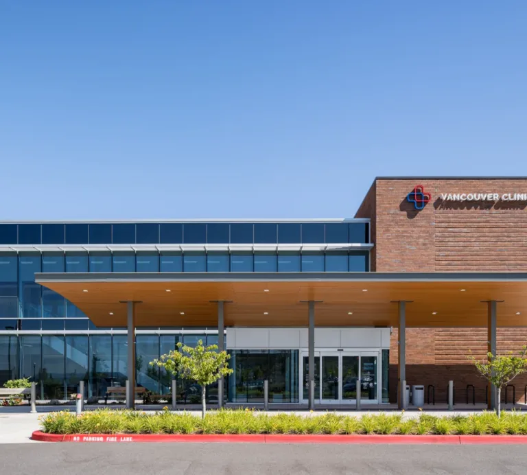 Front view of Vancouver Clinic’s modern building with glass facade and brick sign, a large covered entrance, landscaped grounds, and flags under a clear blue sky.