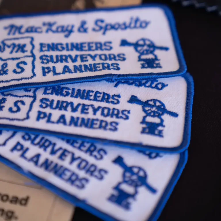 Historical photo of stacked MacKay & Sposito embroidered patches reading Engineers, Surveyors, Planners with a cartoon surveyor icon, resting on a newspaper.