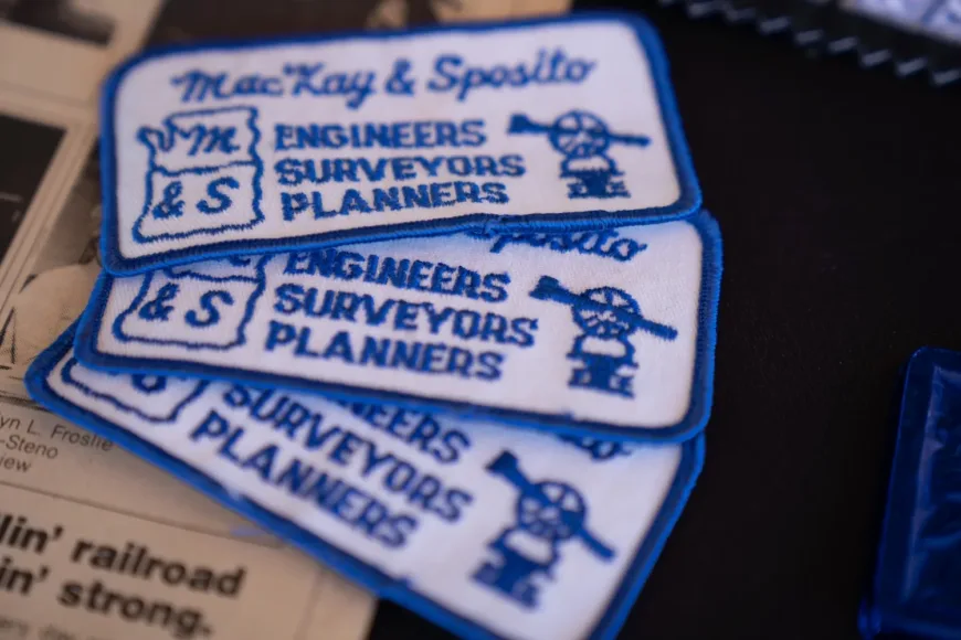 Historical photo of stacked MacKay & Sposito embroidered patches reading Engineers, Surveyors, Planners with a cartoon surveyor icon, resting on a newspaper.