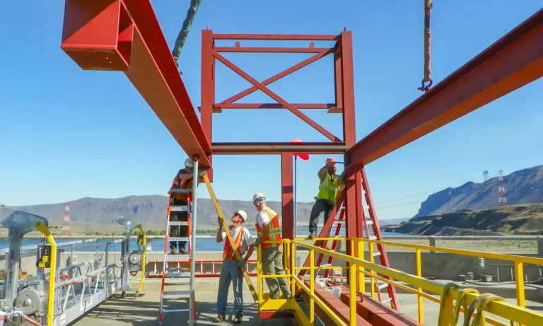 Construction workers in hard hats and safety vests assemble a red steel framework using ladders and a crane on an outdoor industrial site beside a river, with mountains and power lines in the background.