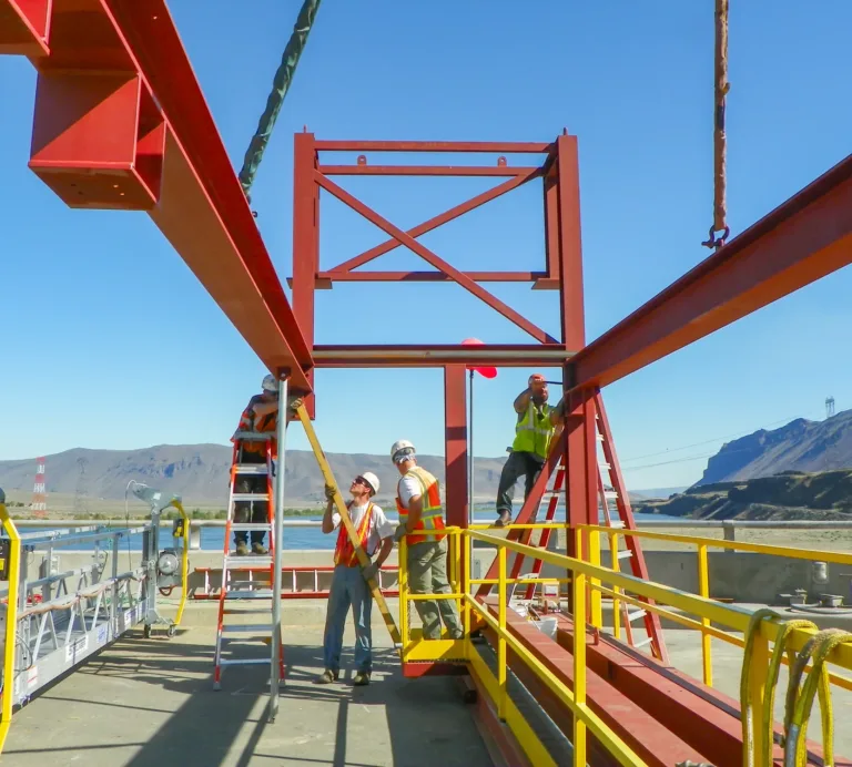 Construction workers in hard hats and safety vests assemble a red steel framework using ladders and a crane on an outdoor industrial site beside a river, with mountains and power lines in the background.