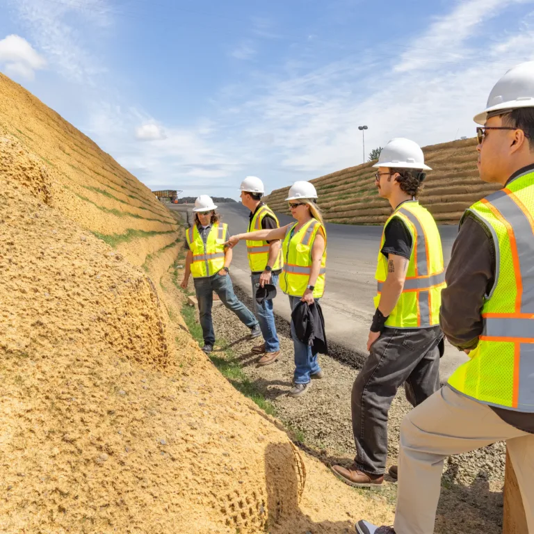 Five construction workers in hard hats and hi-vis vests inspect a stabilized dirt embankment beside a road.