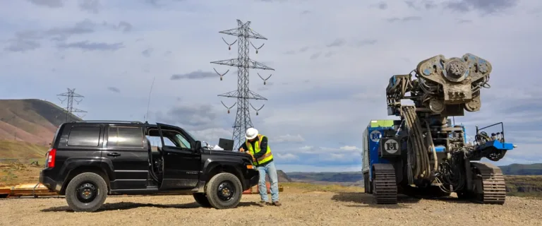 Engineer in safety gear beside a black SUV near a large tracked drilling machine, with power lines and mountains under a cloudy sky.