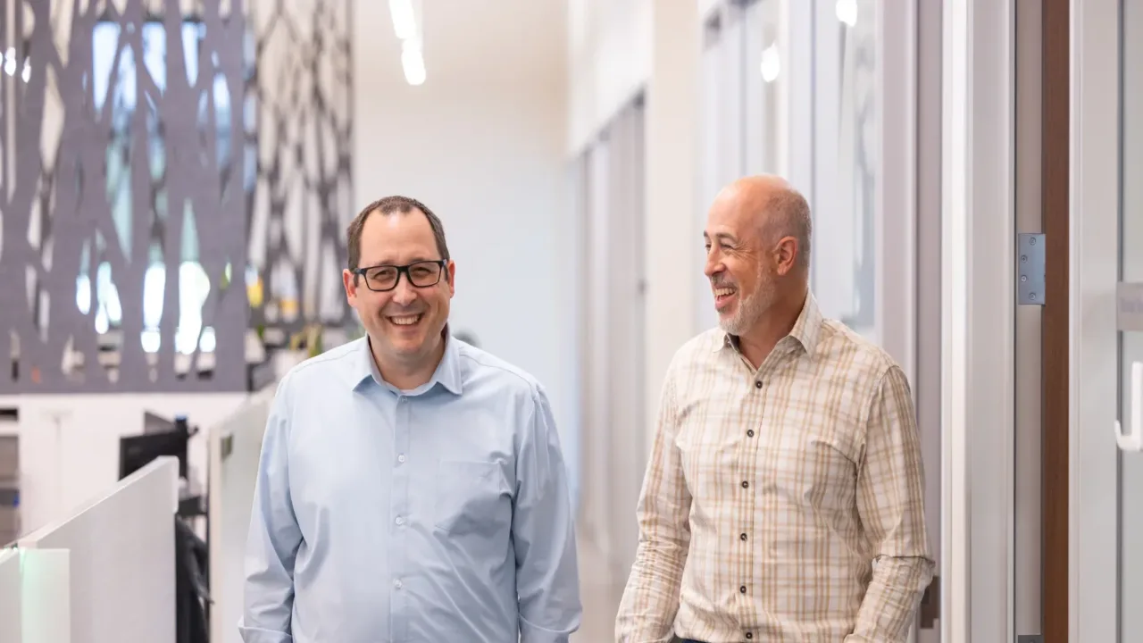 Two MacKay Sposito team members smile while walking down a bright, modern office hallway.