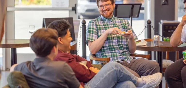MacKay Sposito team members relax and chat in a casual office lounge during a social, with one person smiling and holding a plate of snacks.