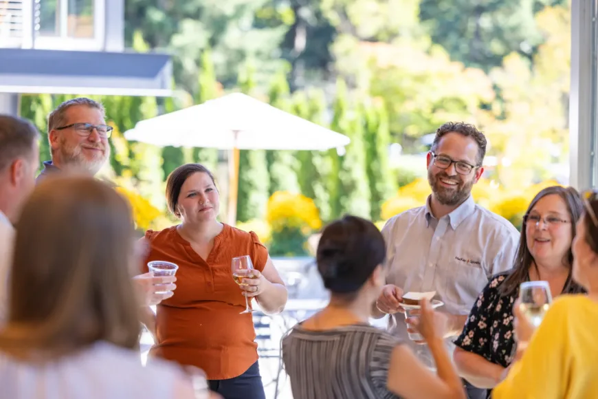 MacKay Sposito team members mingle at an outdoor gathering, smiling and holding drinks on a sunny patio with greenery and umbrellas in the background.